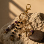 Ariel La Boutique beaded charm keychain with gold cross, dragonfly, and Hamsa hand charms on a textured stone surface.